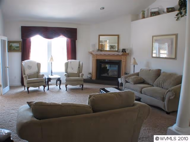 A cozy living room with beige carpet, two beige sofas, two armchairs near a large window with red curtains, a wooden coffee table, a fireplace with a wooden mantel and a mirror above it, and framed artwork on the walls.