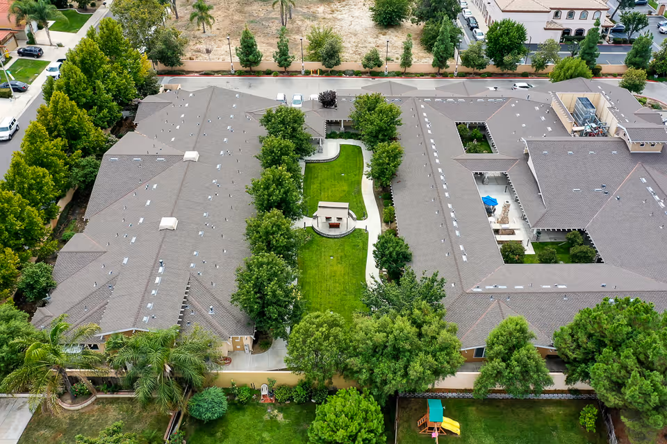 Aerial view of Sunny Rose Senior Living facility showing two large buildings with gray roofs surrounding a green courtyard with trees, pathways, and a small covered seating area. The surrounding area includes trees, a playground with a slide, and nearby streets with parked cars.