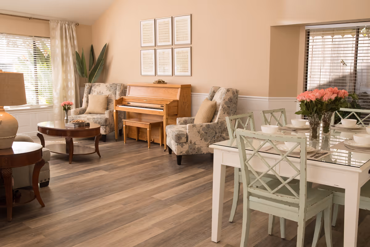 A cozy living and dining area with a wooden piano against the wall, two floral upholstered armchairs with beige cushions, a round wooden coffee table, and a white dining table with four green chairs. The dining table is set with white bowls and plates, and a vase of pink roses is placed in the center. The room has large windows with blinds and light curtains, and wooden flooring.