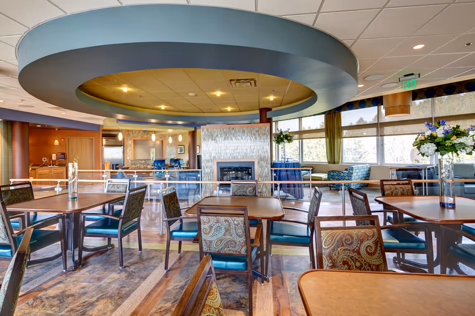 A spacious dining area with multiple tables and chairs featuring patterned upholstery. The room has large windows letting in natural light, a modern circular ceiling design with recessed lighting, and a fireplace with a tiled surround. There are flower arrangements on some tables and a cozy seating area near the windows.