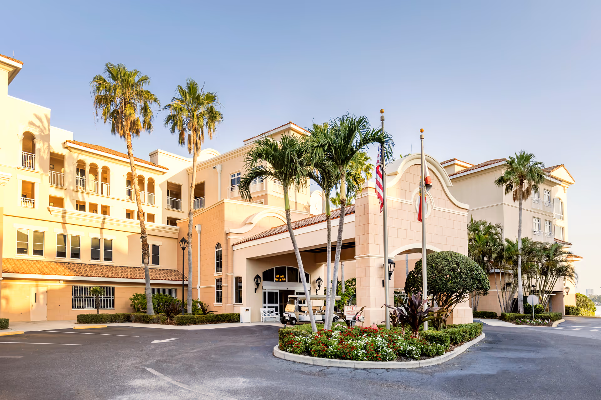 Exterior view of Westminster Shores senior living facility with a driveway, palm trees, and a covered entrance. The building has a light peach color with multiple windows and balconies under a clear blue sky.