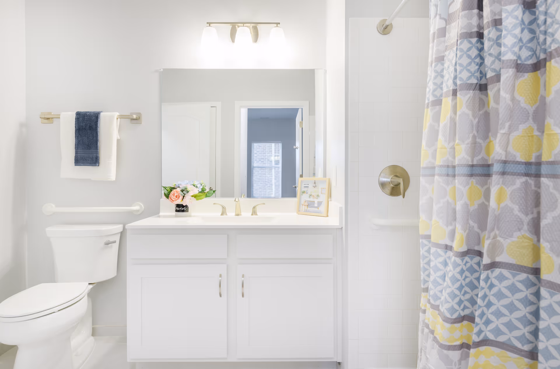 Bright white bathroom featuring a vanity, toilet, grab bars, and a colorful patterned shower curtain.