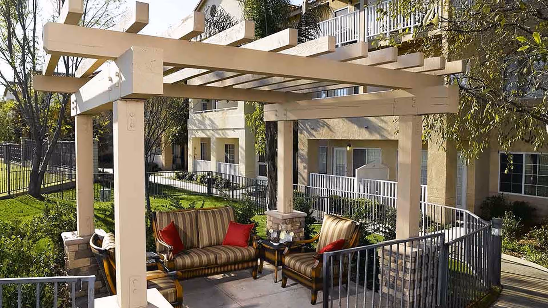 Outdoor seating area at Atria Las Posas featuring a pergola with wooden beams, cushioned chairs and a loveseat with striped upholstery and red pillows, surrounded by greenery and adjacent to the building with balconies and railings.