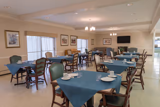 A dining room with multiple tables covered with blue tablecloths, each set with white plates, cups, and glasses. The room has several chairs around the tables, framed artwork on the walls, large windows with curtains, and ceiling lights. A television is mounted on the far wall above a fireplace.
