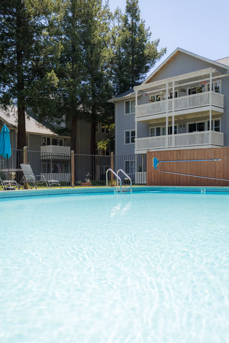 Outdoor swimming pool with clear blue water in front of a multi-story residential building with balconies. There are lounge chairs and a blue umbrella near the pool, and tall trees in the background.