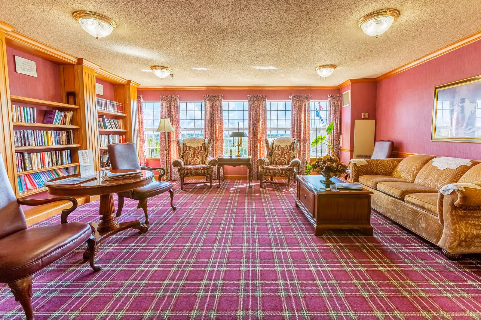 A cozy living room with a purple plaid carpet, two patterned armchairs, a brown sofa, a wooden coffee table with a floral arrangement, a round wooden table with two leather chairs, and a built-in wooden bookshelf filled with books. Large windows with floral curtains let in natural light and offer a view outside.