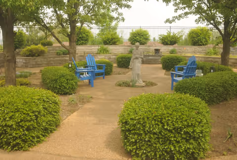 A peaceful outdoor garden area with a circular paved pathway surrounded by neatly trimmed bushes and trees. In the center of the circle is a statue of a person. Four blue Adirondack chairs are arranged around the statue, providing seating in the garden.