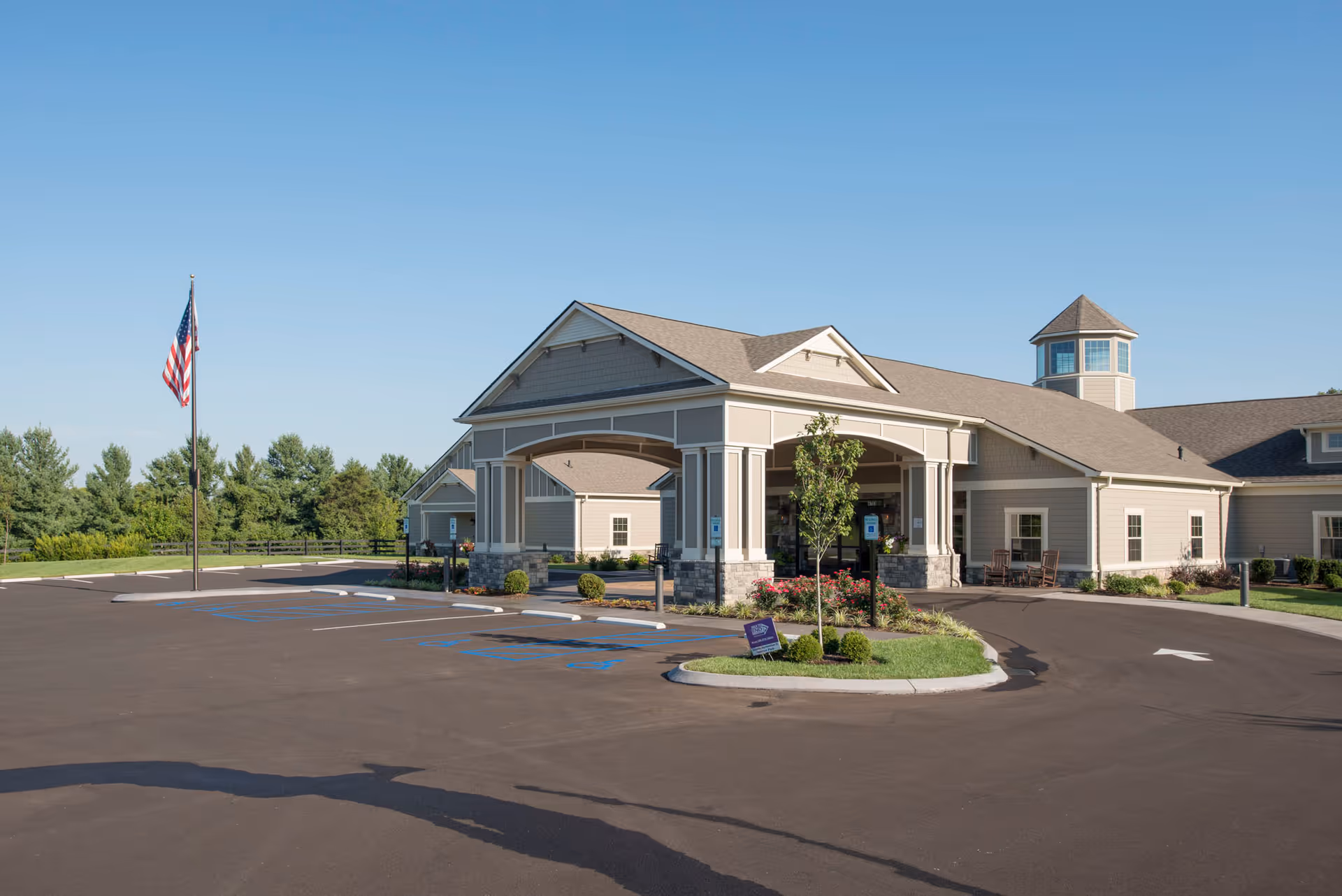 Exterior view of The Lantern at Morning Pointe Alzheimer’s Center of Excellence in Louisville, showing the building entrance with a covered drop-off area, an American flag on a flagpole, parking spaces including handicapped spots, and landscaped greenery under a clear blue sky.