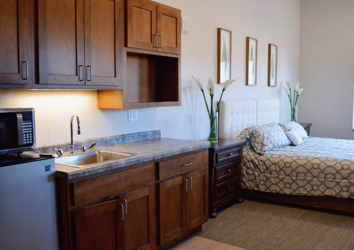 A cozy senior living room featuring a kitchenette with wooden cabinets, a small refrigerator, microwave, and a sink. Adjacent to the kitchenette is a bed with a patterned blue and white bedspread, two nightstands with vases holding white flowers, and three framed botanical prints on the wall.