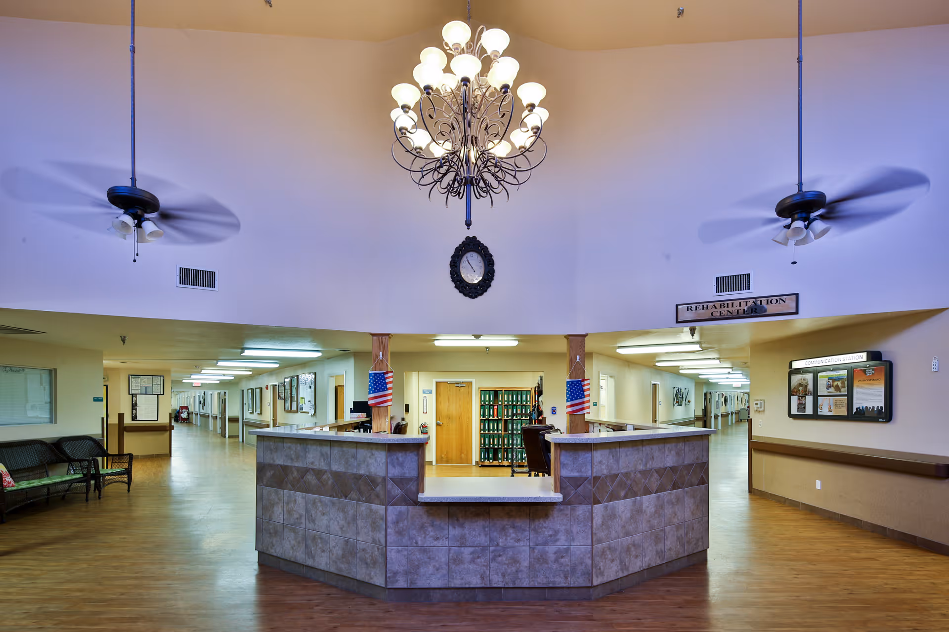 Lobby reception desk of a nursing facility with a central chandelier, two ceiling fans, seating and hallways leading to rooms.