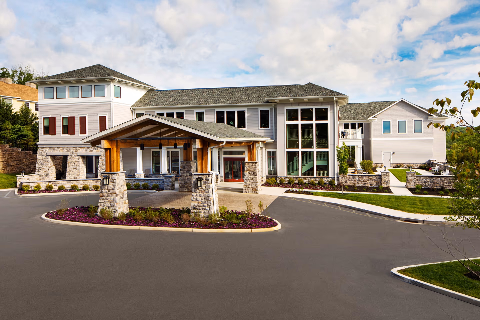 Front exterior view of Ridgefield Station Senior Living facility showing a large building with multiple windows, a covered entrance supported by stone pillars, landscaped flower beds, and a paved driveway under a partly cloudy sky.