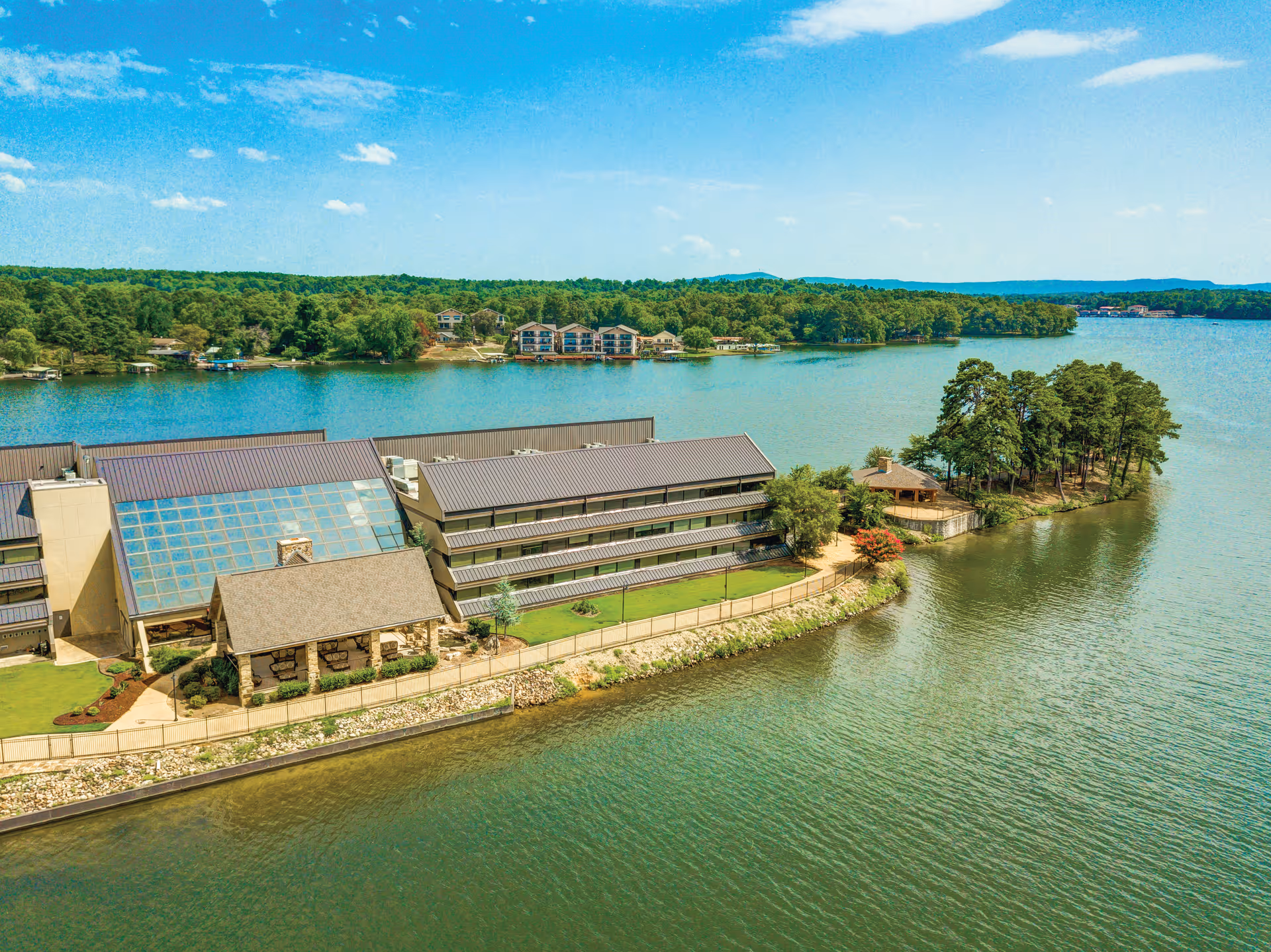 Aerial view of The Atrium at Serenity Pointe, a large building complex situated on a waterfront with a lake surrounding it. The building has multiple floors with balconies, a glass atrium, and a small gazebo-like structure near the water. Trees and greenery surround the area, with more buildings visible across the lake under a clear blue sky.