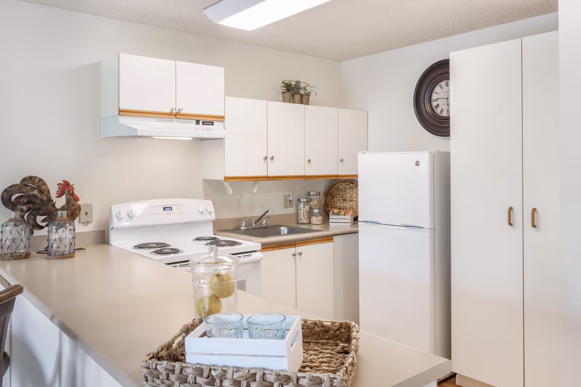 A clean and bright kitchen with white cabinets, a white electric stove, a white refrigerator, and a countertop with decorative items including a wicker tray holding glasses and a jar with green apples. A large wall clock is mounted above the refrigerator.