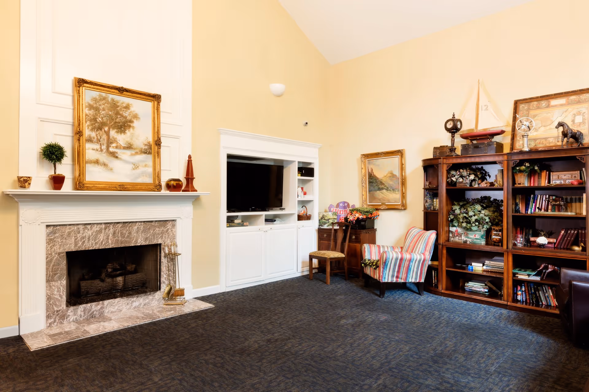 A cozy living room with a marble fireplace on the left, a white built-in entertainment center with a TV, a striped armchair, a wooden chair, and a wooden bookshelf filled with books and decorative items. The walls are painted light yellow and there are framed paintings hanging on the walls.