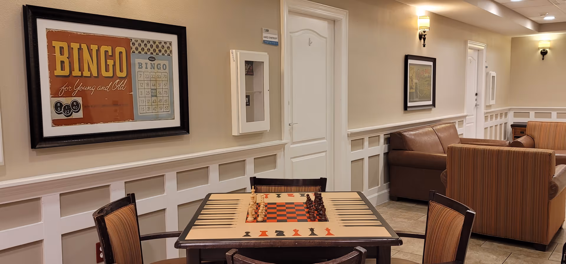 A cozy common area in a senior living facility featuring a game table with chess and backgammon, surrounded by four chairs. The walls are beige with white wainscoting, decorated with framed artwork including a bingo-themed poster. There are two brown sofas and a striped armchair in the background, with soft wall lighting illuminating the space.