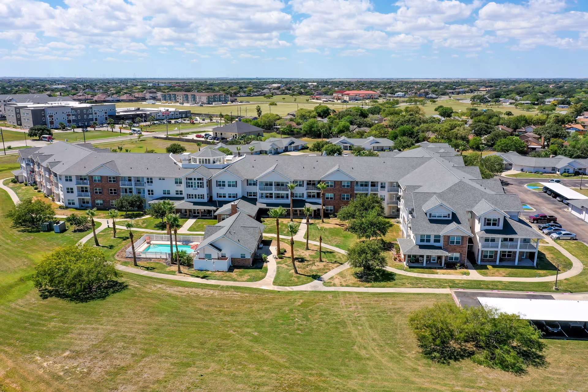 Aerial view of a large senior living facility with multiple connected buildings, surrounded by green lawns and trees. The facility includes a swimming pool area with palm trees and paved walkways. In the background, there are residential neighborhoods and other buildings under a partly cloudy sky.