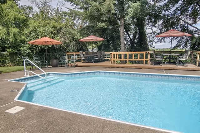 Outdoor swimming pool with clear blue water, surrounded by a concrete deck. In the background, there is a wooden deck with tables, chairs, and red umbrellas. Trees and greenery surround the area.