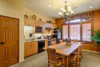 Dining area with wooden table, chairs, chandelier, and a kitchen in the background.