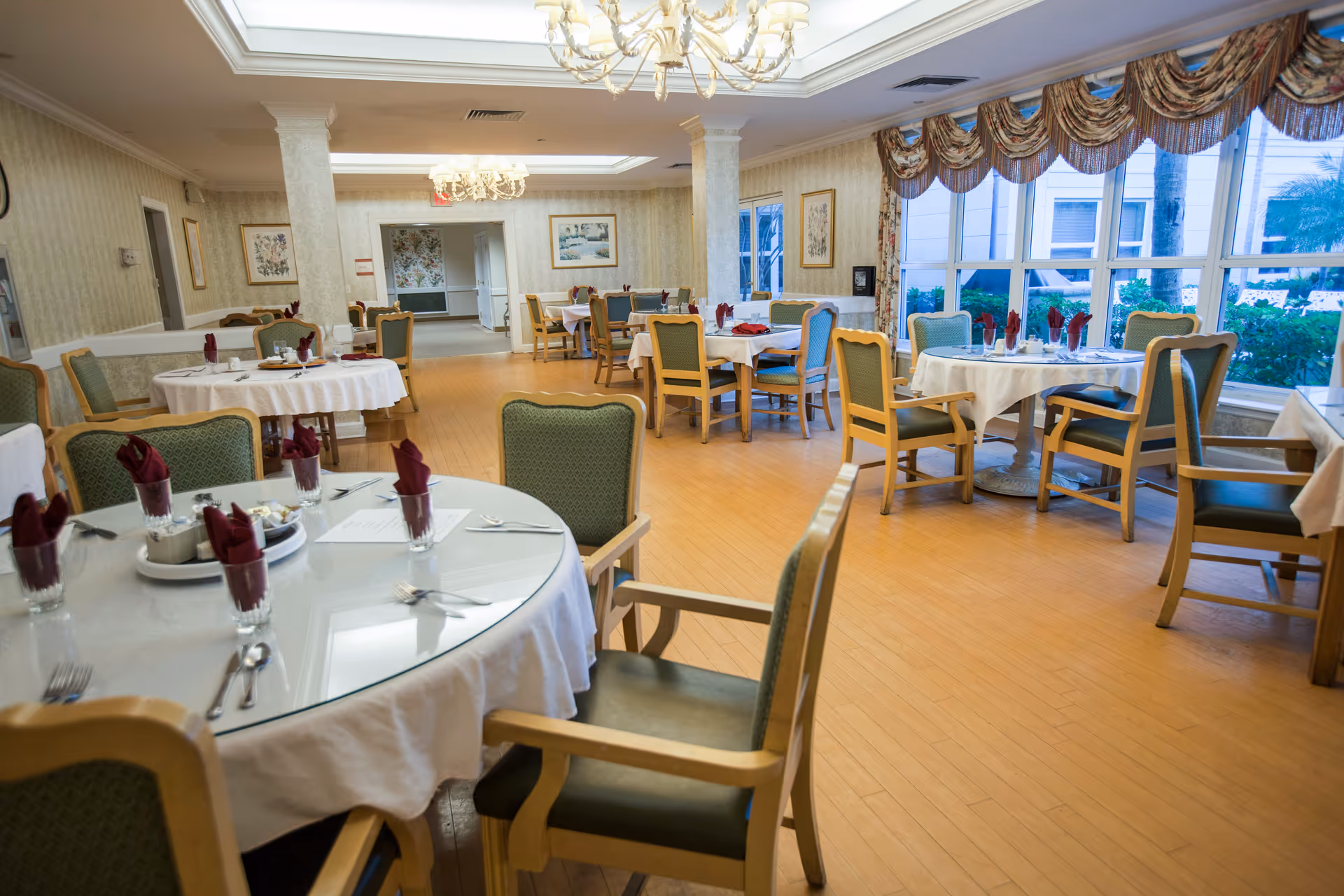 A spacious dining room with multiple round tables covered with white tablecloths, each set with glasses containing folded burgundy napkins, silverware, and plates. The room features wooden chairs with green upholstery, large windows with decorative valances, chandeliers hanging from the ceiling, and framed artwork on the walls. The floor is light wood, and greenery is visible outside the windows.