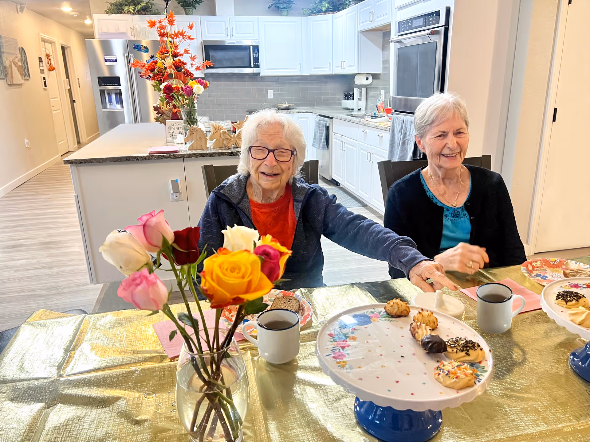 Two elderly women sitting at a dining table in a bright kitchen. The table is covered with a gold tablecloth and has a vase with colorful roses and a plate with assorted cookies. The kitchen in the background has white cabinets, a refrigerator, microwave, and oven. Both women are smiling and appear to be enjoying their time together.