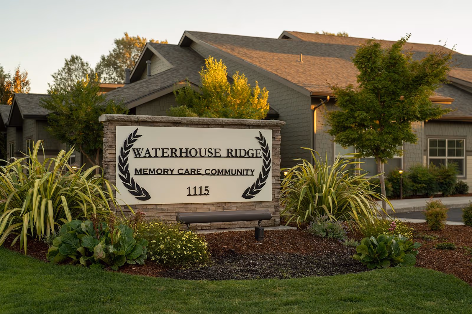 Outdoor view of the entrance sign for Waterhouse Ridge Memory Care Community, surrounded by landscaped greenery and plants, with a building in the background.