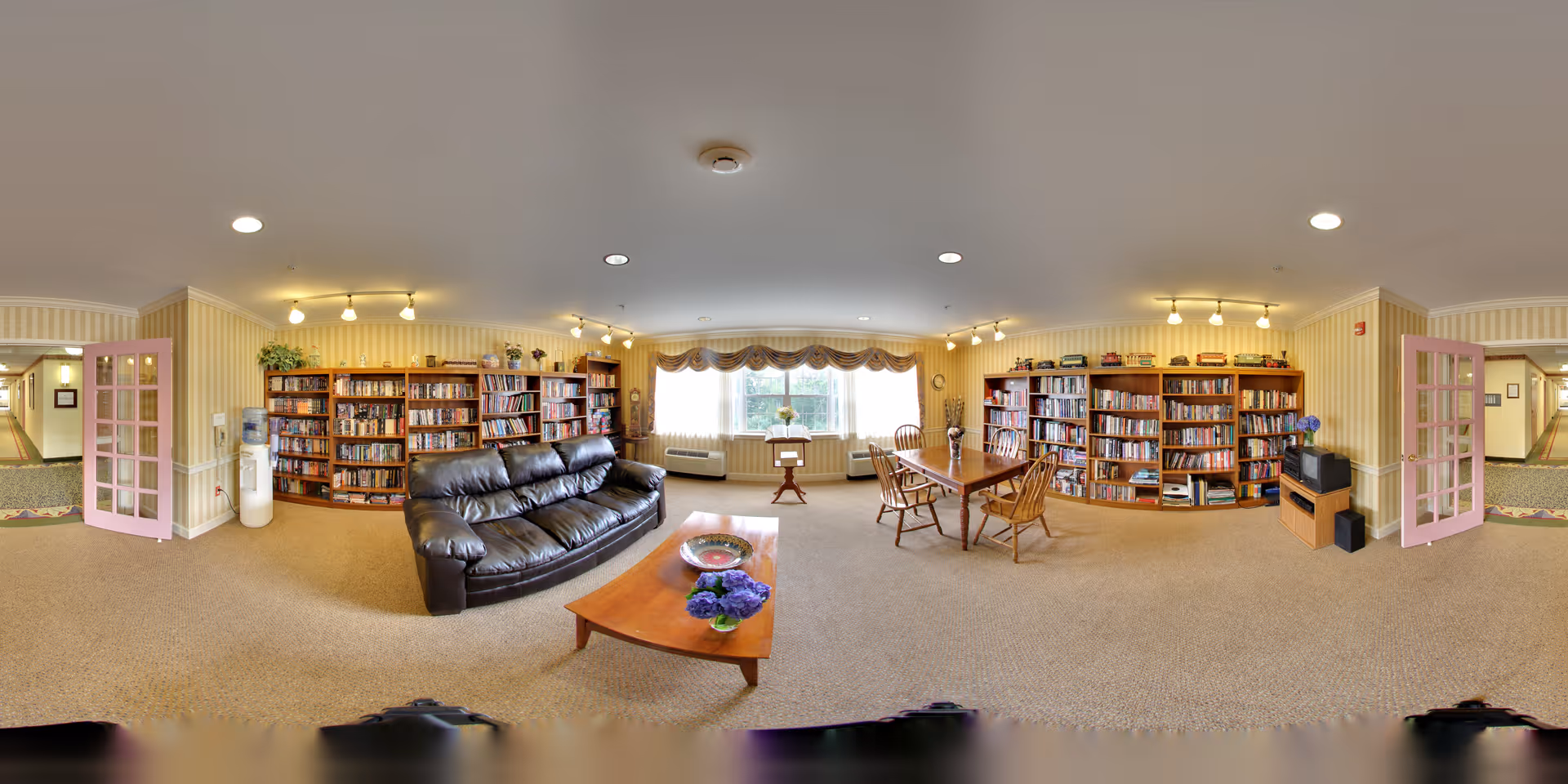 Spacious senior living common room with bookshelves, a leather sofa, coffee table and a table with chairs under soft overhead lighting.