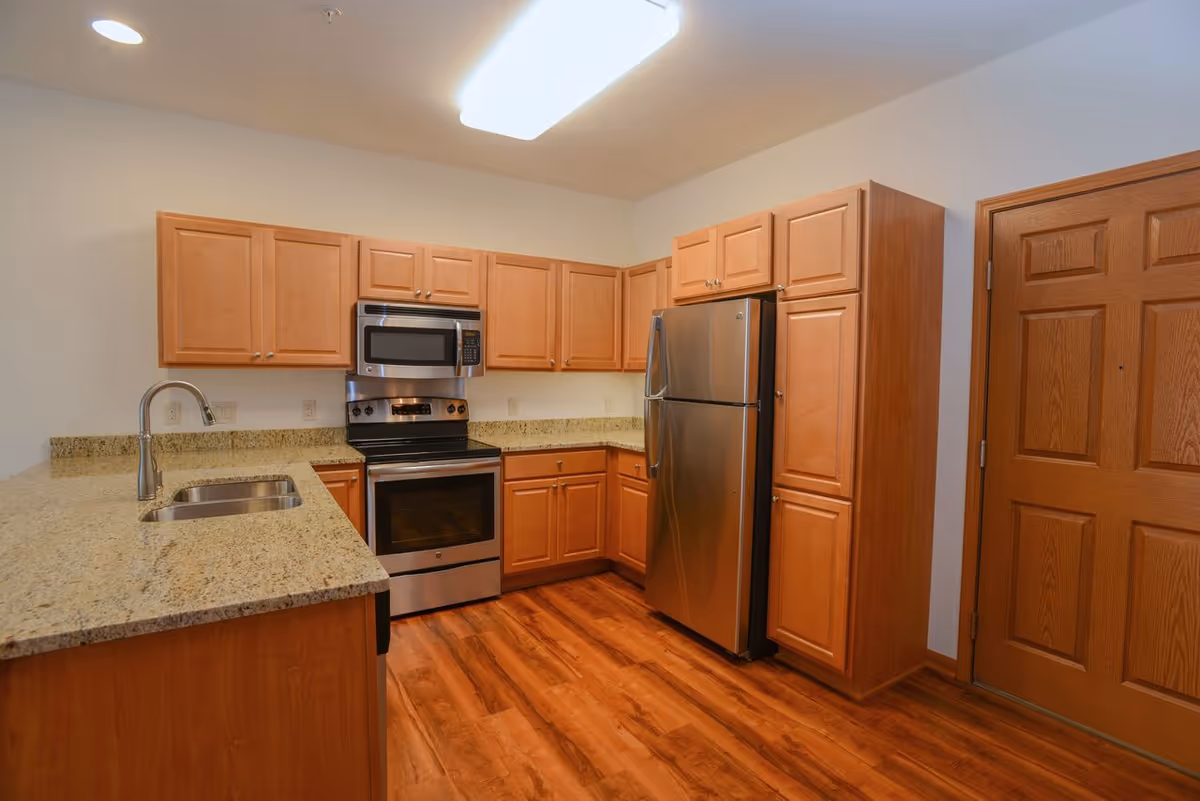 A modern kitchen with wooden cabinets, granite countertops, a stainless steel refrigerator, oven, and microwave. The kitchen has a double sink with a curved faucet and wood flooring. There is a wooden door to the right side of the kitchen.