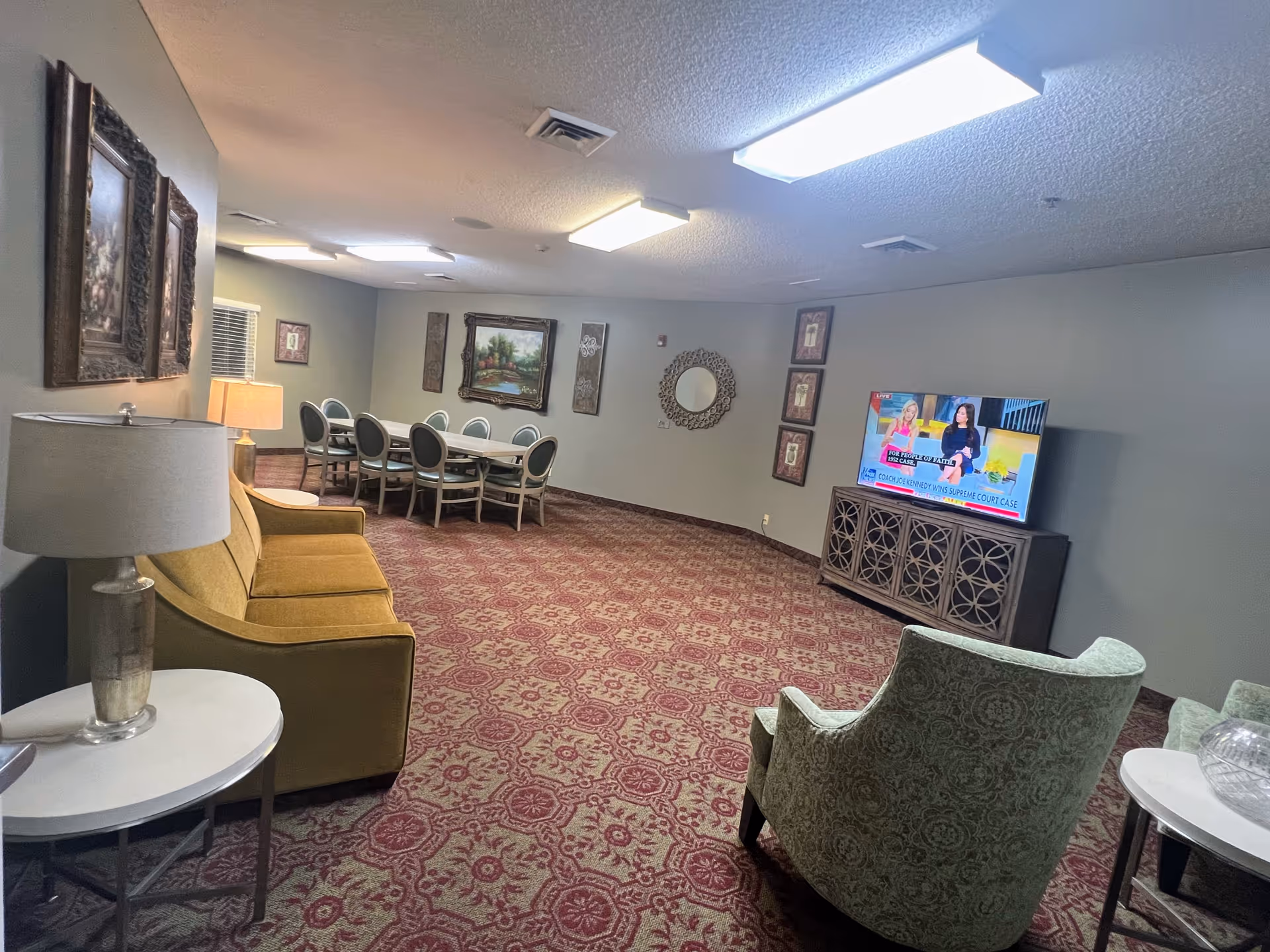 A cozy common area in a senior living facility featuring a mustard yellow sofa, a green patterned armchair, a flat-screen TV on a decorative wooden stand, a dining table with eight chairs, framed artwork on the walls, and a patterned red carpet.