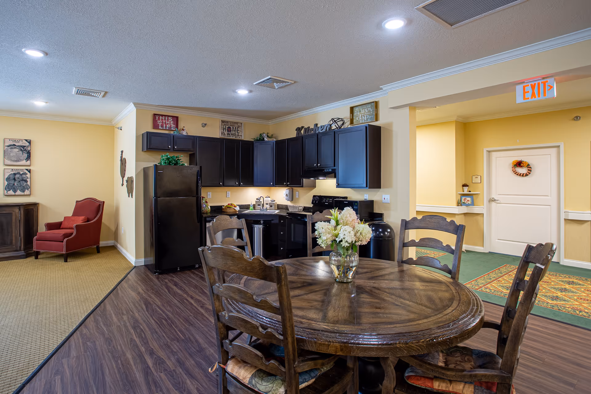 Interior view of a senior living facility kitchen and dining area with a round wooden table and four chairs, a black refrigerator, dark wood cabinets, a stove, and a sink. There is a red armchair and a wooden cabinet against a yellow wall in the background. The floor transitions from wood to carpet, and there is an exit sign above a hallway door decorated with a wreath.