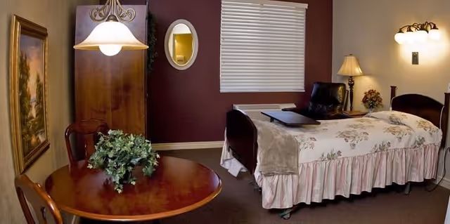 A cozy bedroom in a senior living facility featuring a single bed with floral bedding and a beige throw blanket. Next to the bed is a black recliner chair and a small table with a floral arrangement. The room has a round wooden table with two chairs and a green plant centerpiece. There is a wooden wardrobe, a framed painting on the wall, an oval mirror, and a window with closed blinds. The lighting includes a hanging lamp above the table, a wall-mounted light fixture, and a table lamp.
