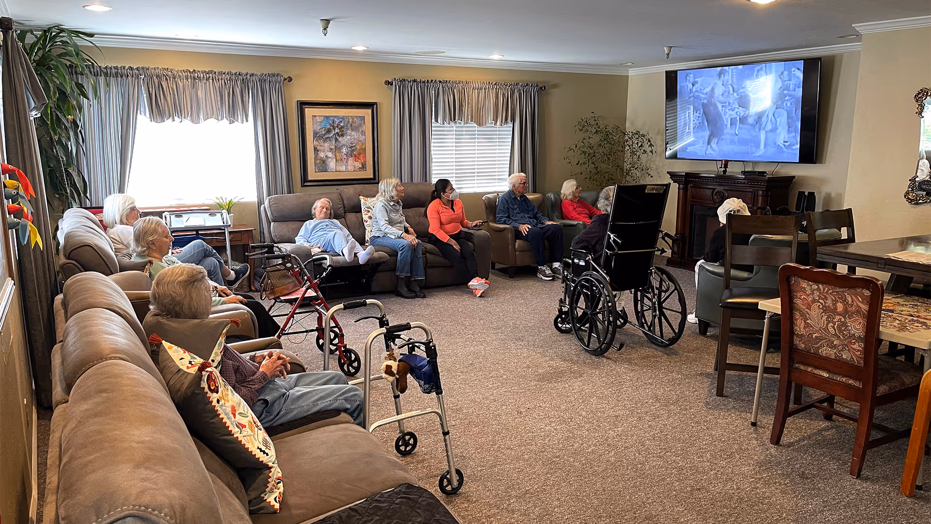 A group of elderly people sitting in a living room area of an elderly care facility. They are seated on sofas and chairs arranged in a semi-circle facing a television mounted on the wall. Some walkers and a wheelchair are visible in the room. The room has beige walls, carpeted floor, and windows with curtains allowing natural light in.