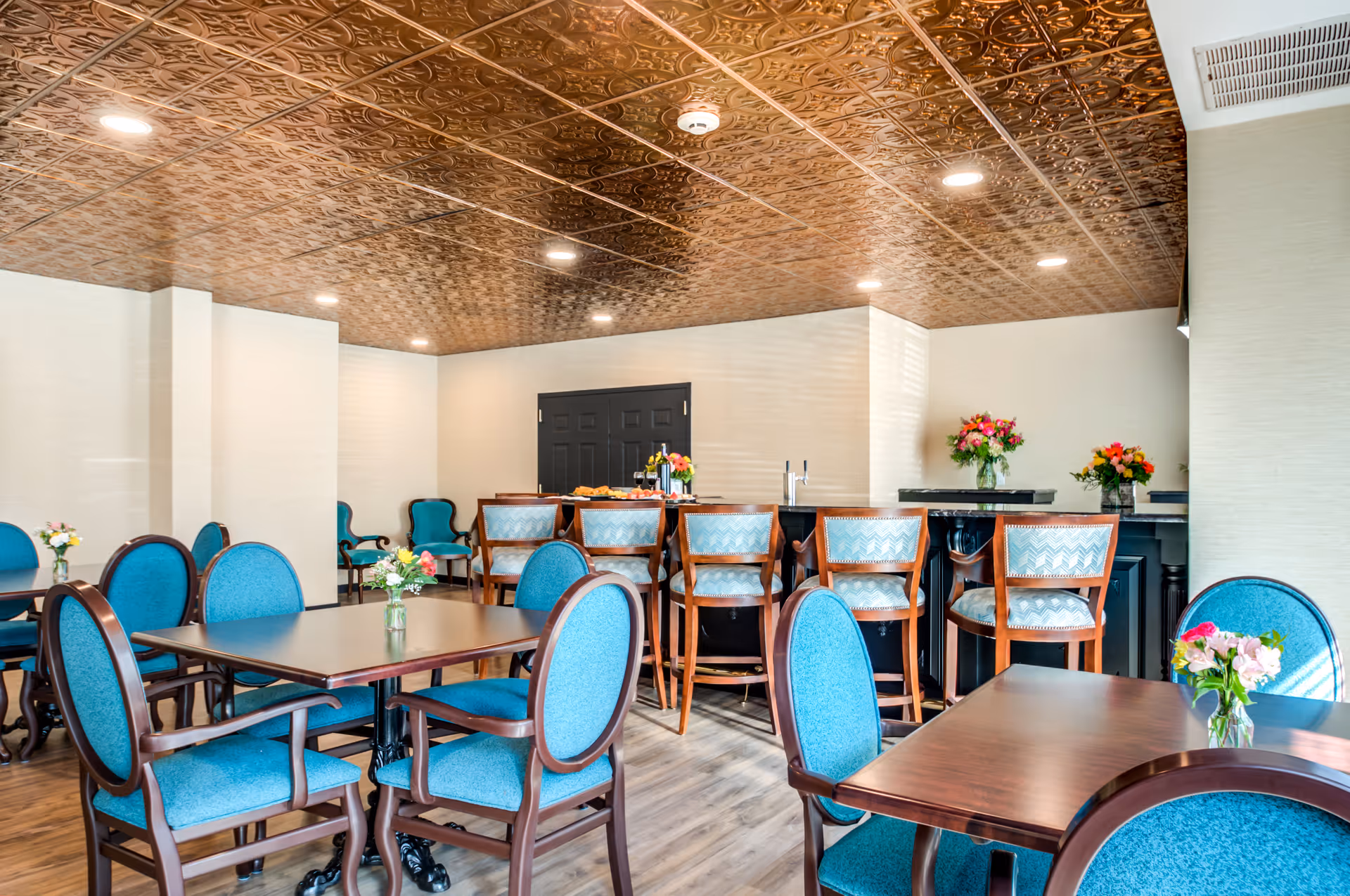 A dining room with several wooden tables and blue upholstered chairs. The room features a decorative copper-colored ceiling with recessed lighting. There is a bar area with high chairs and flower arrangements on the tables and bar counter.