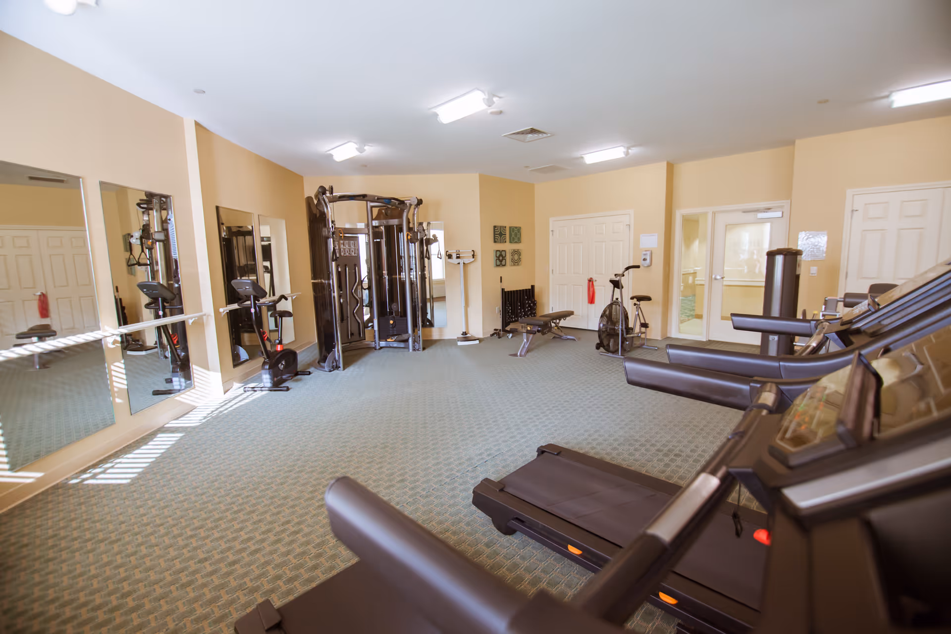 A fitness room with exercise equipment including treadmills, stationary bikes, a multi-functional weight machine, and a bench. The room has beige walls, green patterned carpet, large mirrors on one wall, and bright overhead lighting.
