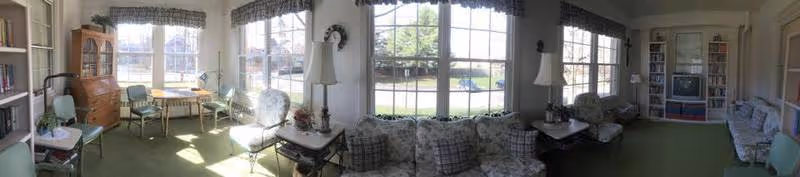 Panoramic view of a sunlit living room with large windows, multiple seating areas including cushioned chairs and sofas, side tables with lamps, a wooden cabinet, and a television. The room has green carpeting and is decorated with curtains and various small decorative items.