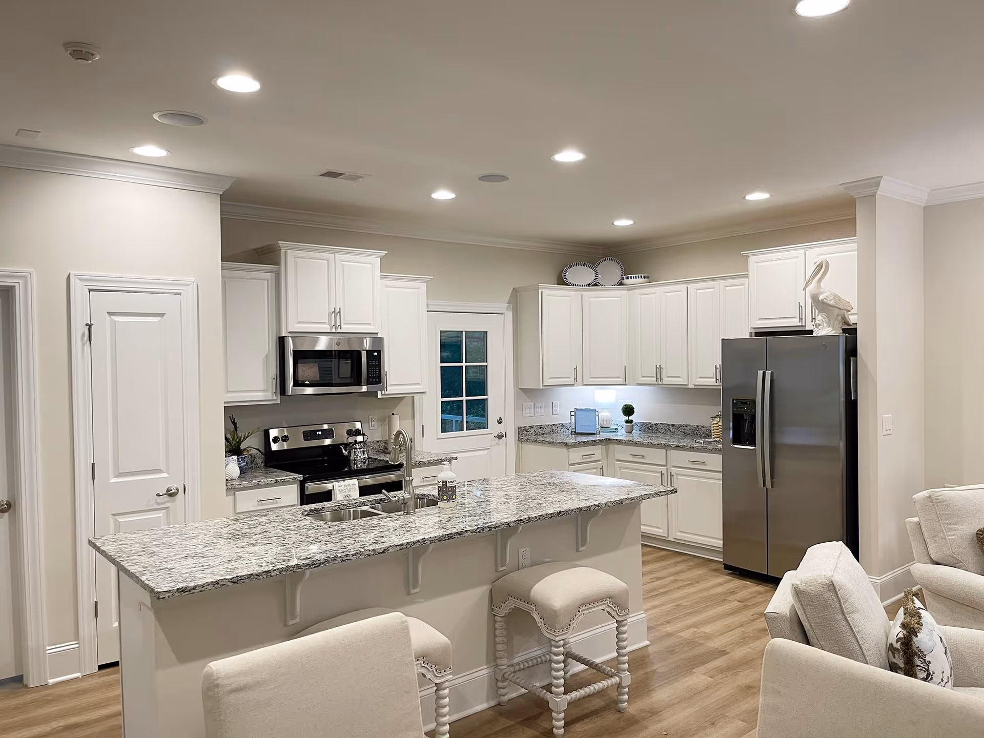 A modern kitchen with white cabinets, granite countertops, and stainless steel appliances including a refrigerator, microwave, and stove. There is a kitchen island with two upholstered stools, and the floor is light wood. The kitchen opens into a living area with beige upholstered chairs.