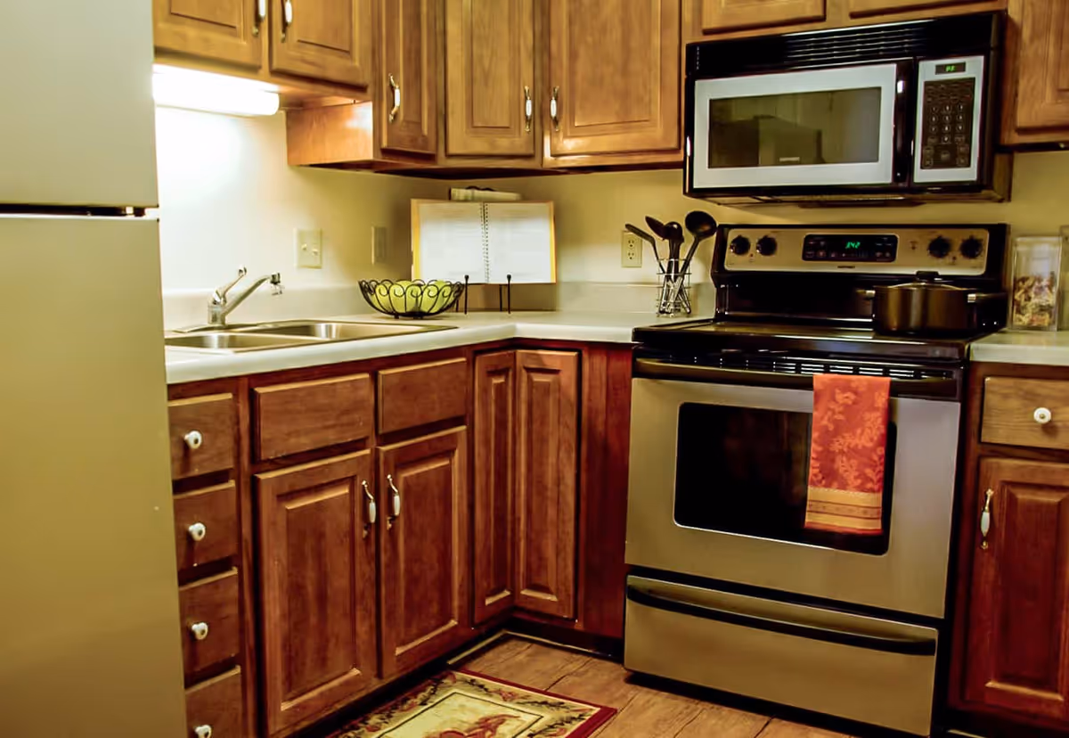 A kitchen with wooden cabinets, a stainless steel stove with a red towel hanging on the handle, a microwave above the stove, a double sink, a refrigerator, and a bowl of green apples on the countertop.