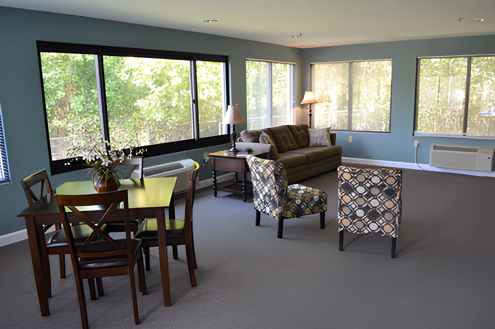 A bright and spacious living room area with large windows letting in natural light. The room features a brown sofa, two patterned armchairs, a wooden dining table with four chairs, and two lamps providing warm lighting. The walls are painted blue and the floor is carpeted in gray.