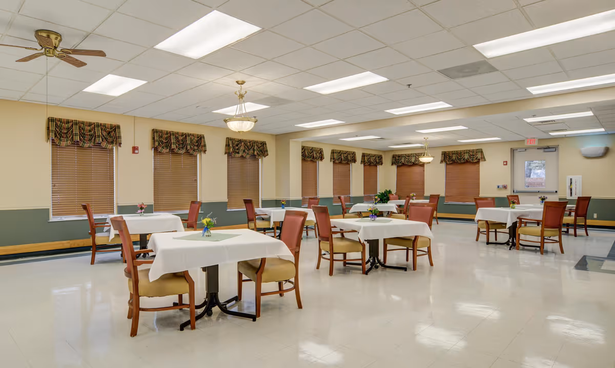 A spacious dining room with multiple square tables covered with white tablecloths, each surrounded by wooden chairs with cushioned seats. The room has several windows with brown blinds and patterned valances. The ceiling features fluorescent lights, a ceiling fan, and chandeliers. Small flower arrangements are placed on each table.