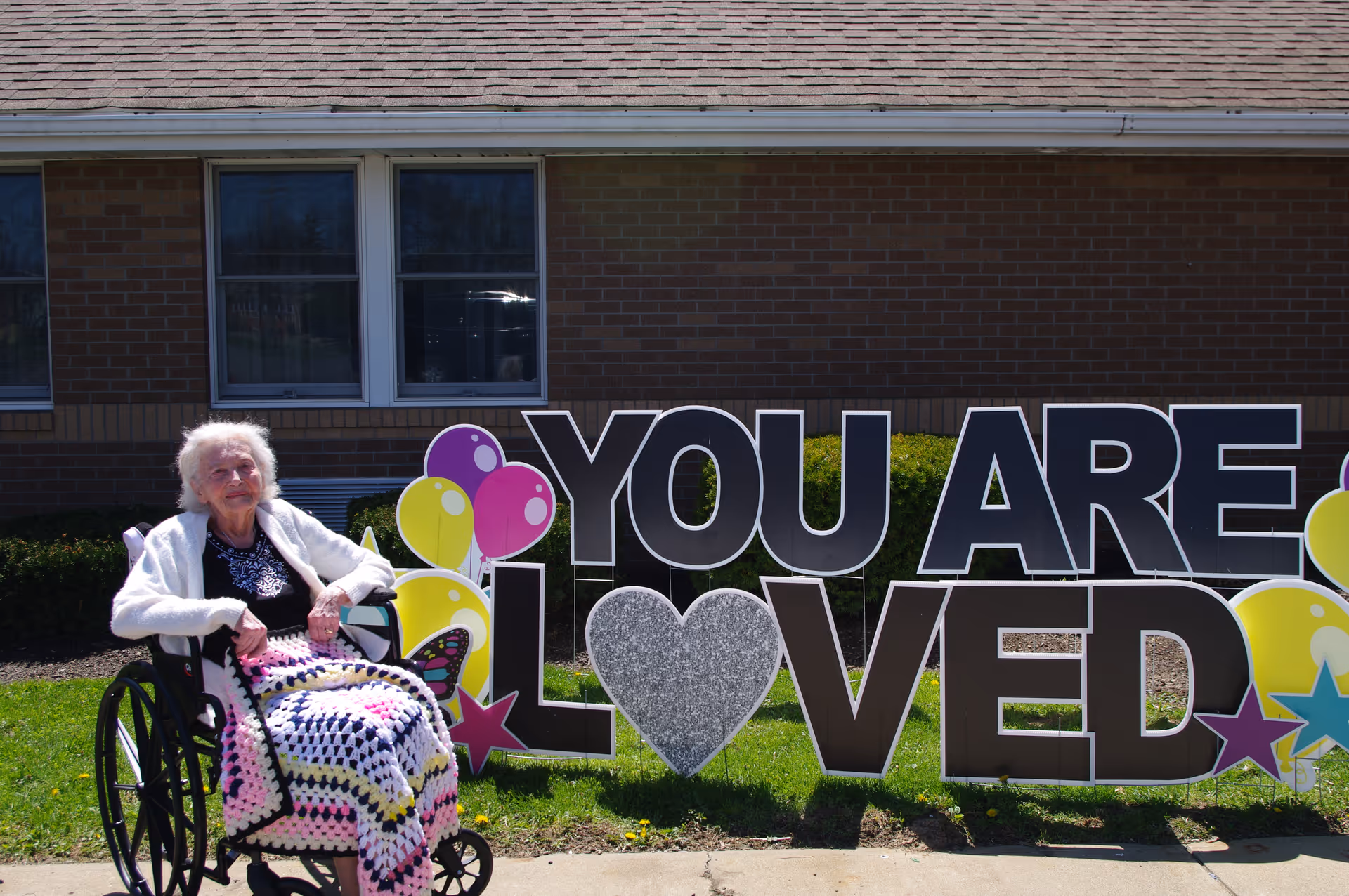 An elderly woman sitting in a wheelchair outdoors in front of a brick building. She is covered with a colorful crocheted blanket and is positioned next to a large decorative sign that reads 'YOU ARE LOVED' with balloons and stars around the text.