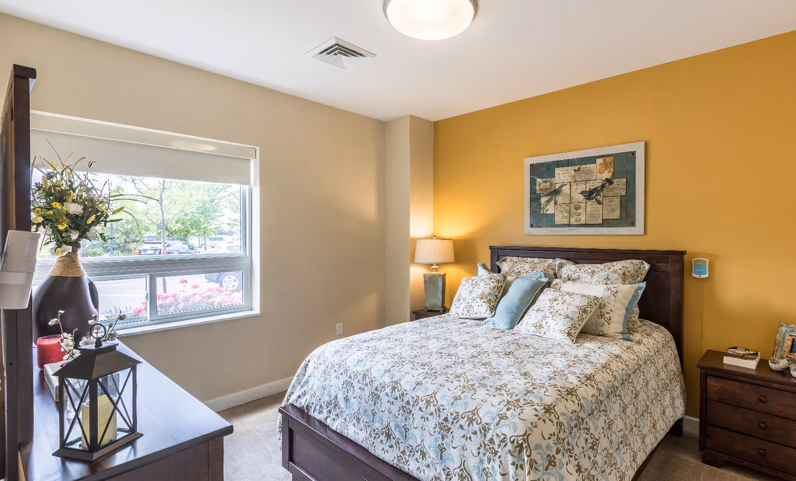 A cozy bedroom with a large bed covered in a floral patterned comforter and multiple pillows. The room has a mustard yellow accent wall behind the bed, a bedside table with a lamp, and a dresser with decorative items including a vase with flowers and a lantern. A window with a white roller shade lets in natural light and shows a view of trees and parked cars outside.