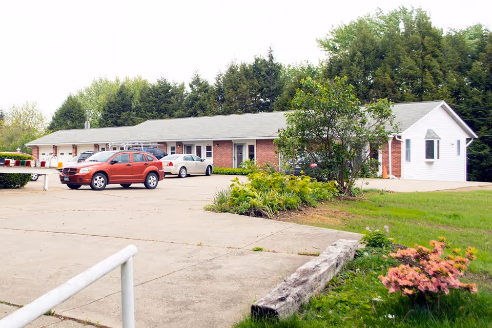 Single-story brick and white retirement home building with parked cars, a driveway, and landscaped front lawn.
