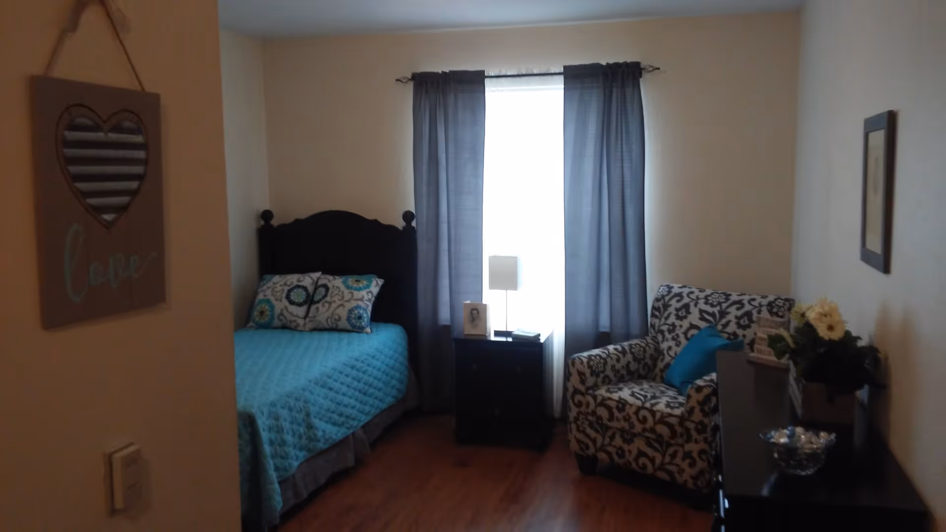 A cozy bedroom with a single bed covered in a turquoise quilt and decorative pillows. Next to the bed is a small black nightstand with a lamp and a framed photo. A window with dark curtains is behind the nightstand. To the right, there is a patterned armchair with a blue cushion and a dark dresser with a flower arrangement and a bowl on top. A wall decoration with a heart and the word 'love' hangs on the left wall.