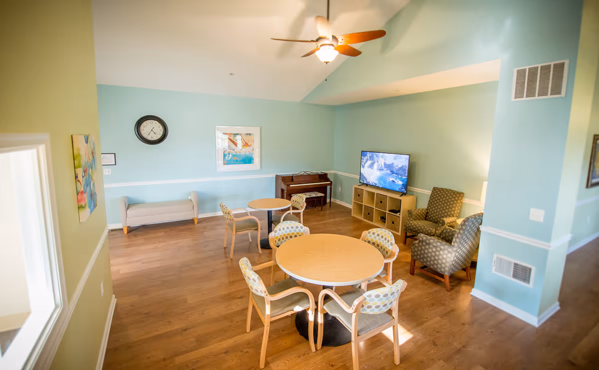 Bright communal living room with round tables and chairs, armchairs, a TV, and a piano on wood floors.