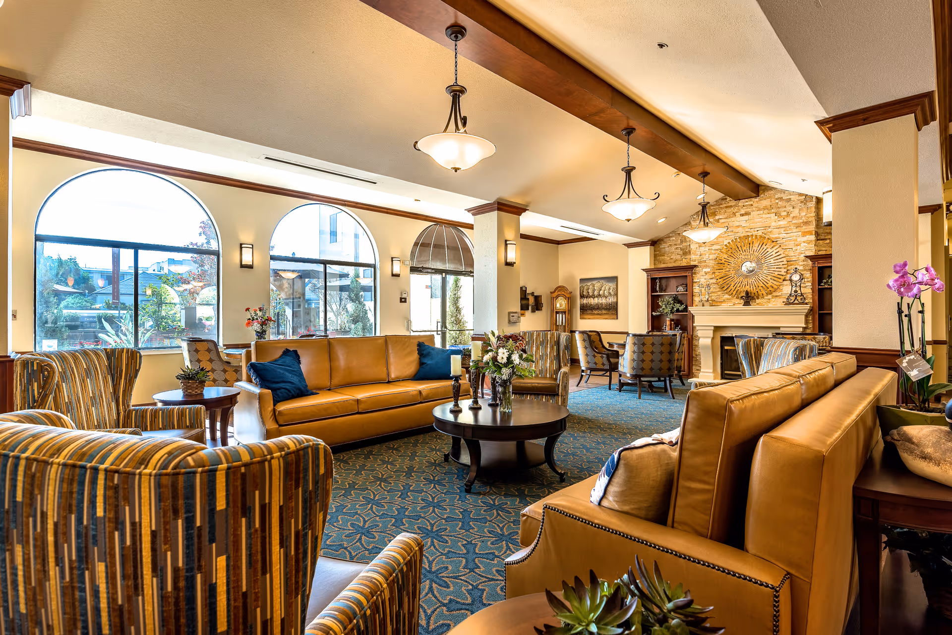 Spacious sunlit lounge in a senior living facility with leather sofas, patterned armchairs, coffee tables, a fireplace, and large arched windows.