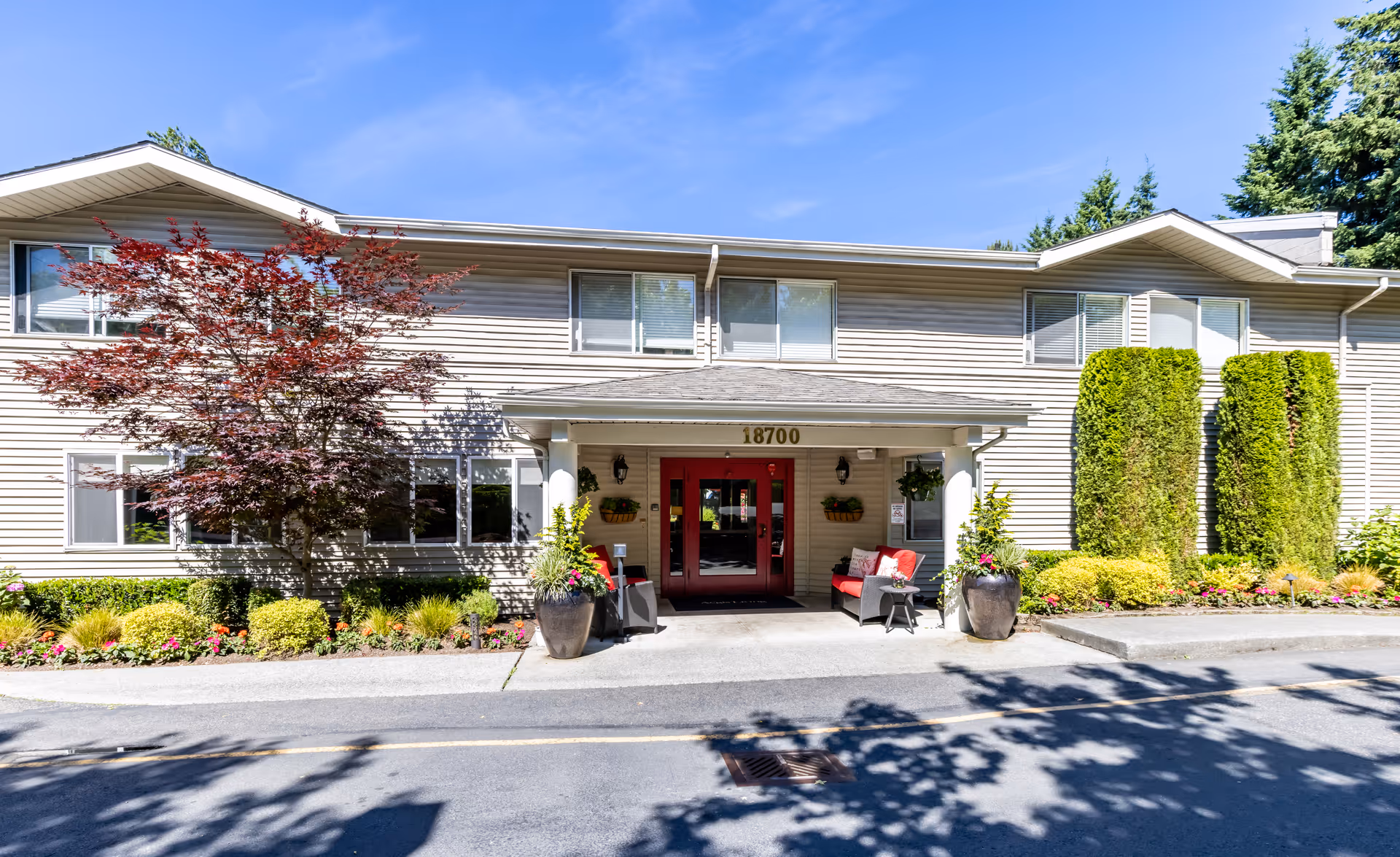 Exterior front entrance of a senior living building with red double doors, potted plants, and landscaped shrubs under a blue sky.