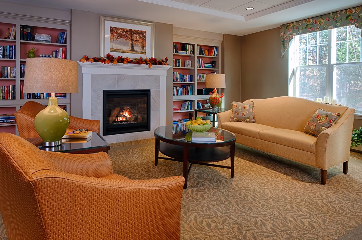 A cozy living room with a beige sofa and two orange armchairs arranged around a round glass coffee table. The room features a fireplace with a lit fire, decorated with autumn leaves and a framed picture above it. Built-in bookshelves filled with books flank the fireplace. A large window with floral valance allows natural light to brighten the space.