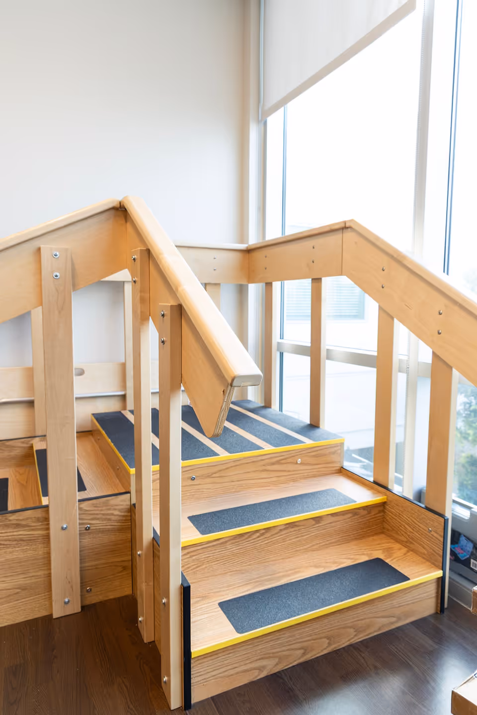 A small wooden staircase with handrails on both sides and non-slip black strips on each step, located next to large windows with natural light coming through.