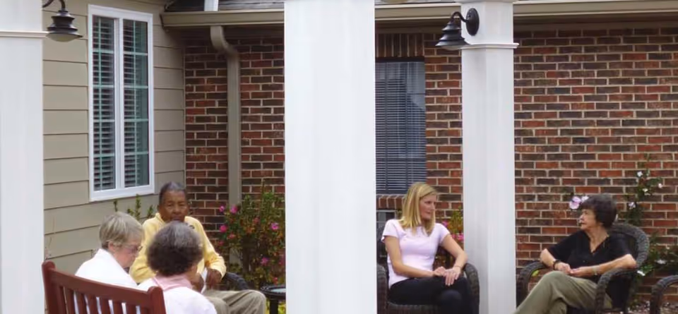 Four elderly people and one younger woman sitting and conversing in an outdoor patio area with brick walls and white pillars, surrounded by plants and flowers.
