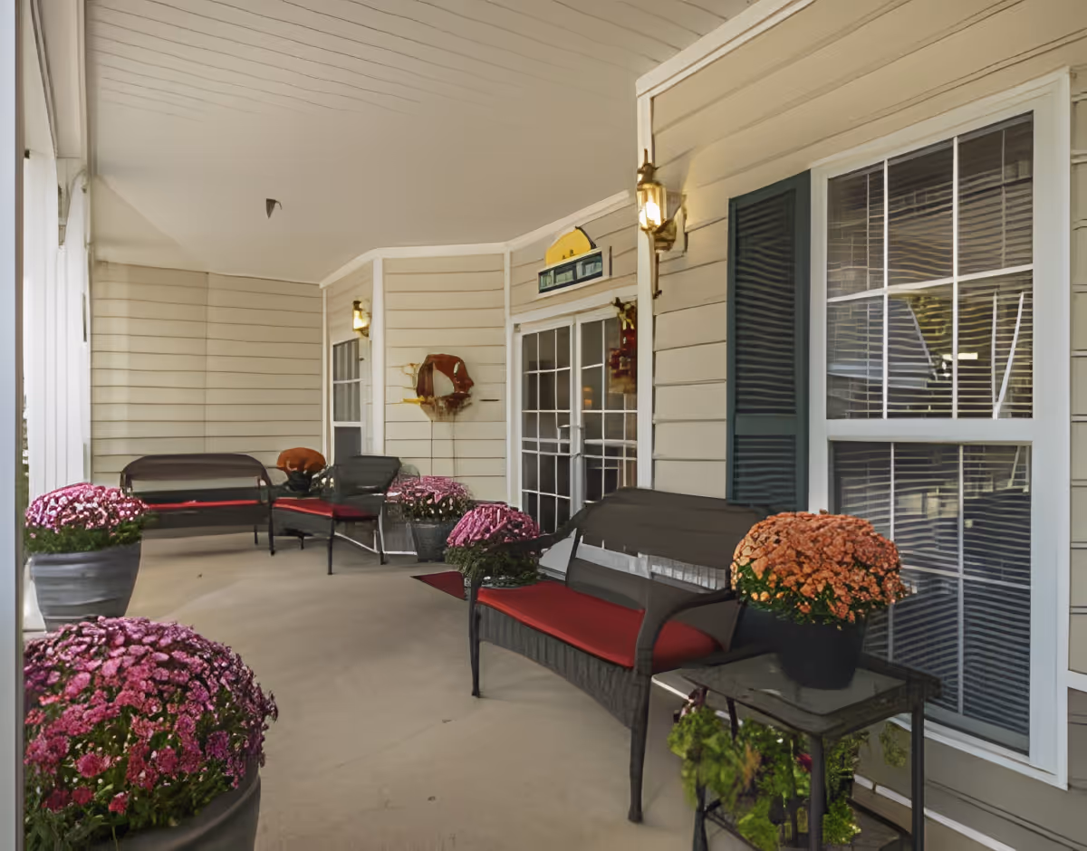 Covered outdoor porch area with beige siding walls, several black metal benches with red cushions, potted flowers in shades of pink and orange, a glass-top side table, and two wall-mounted lantern-style lights near a set of white-framed glass doors and windows.