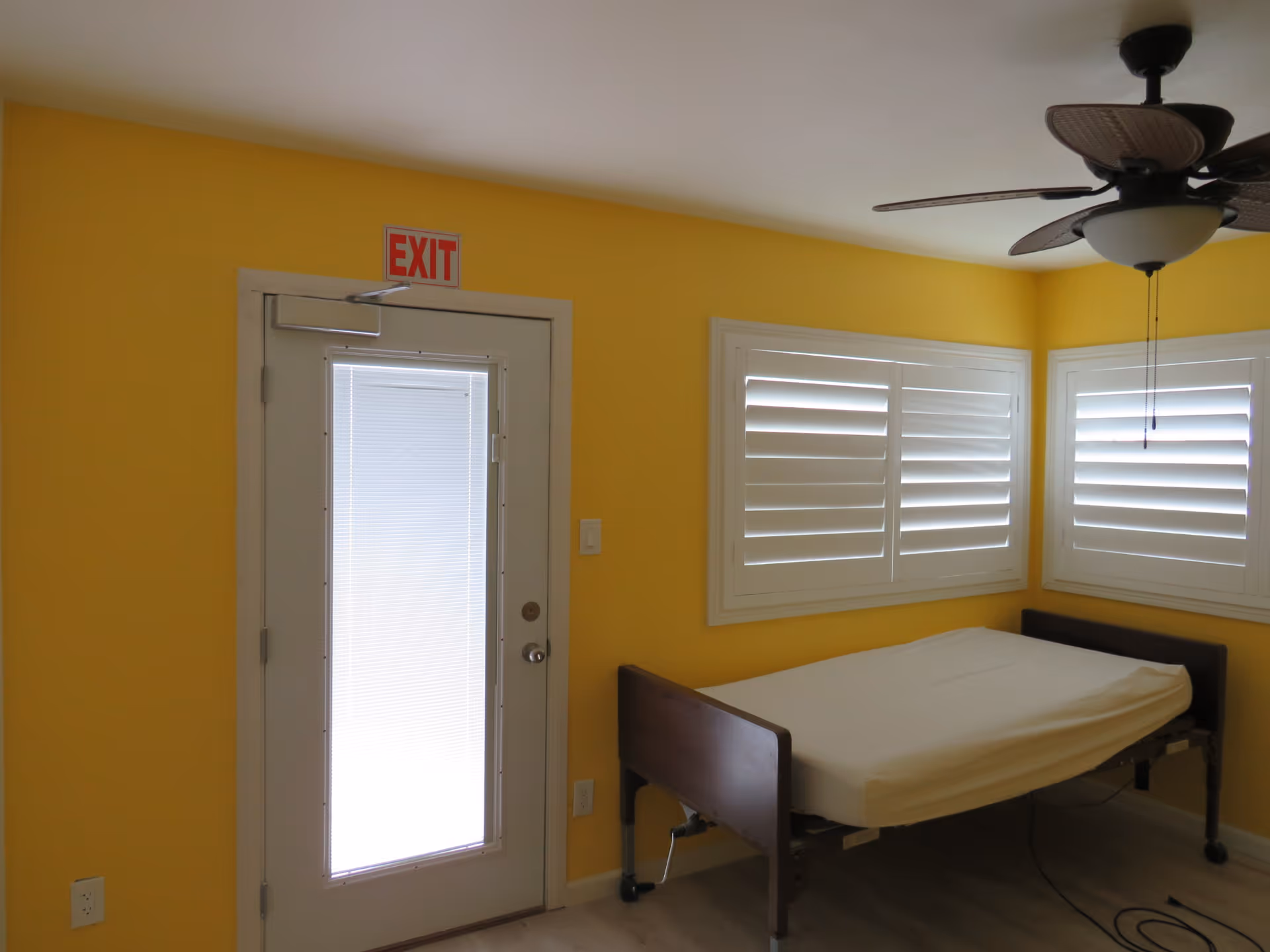 A small room with bright yellow walls featuring a single hospital-style bed with a white mattress. There are two windows with white plantation shutters on the adjacent walls. A ceiling fan with dark blades is mounted on the white ceiling. A door with a large frosted glass panel and an illuminated red EXIT sign above it is on the left side of the room.